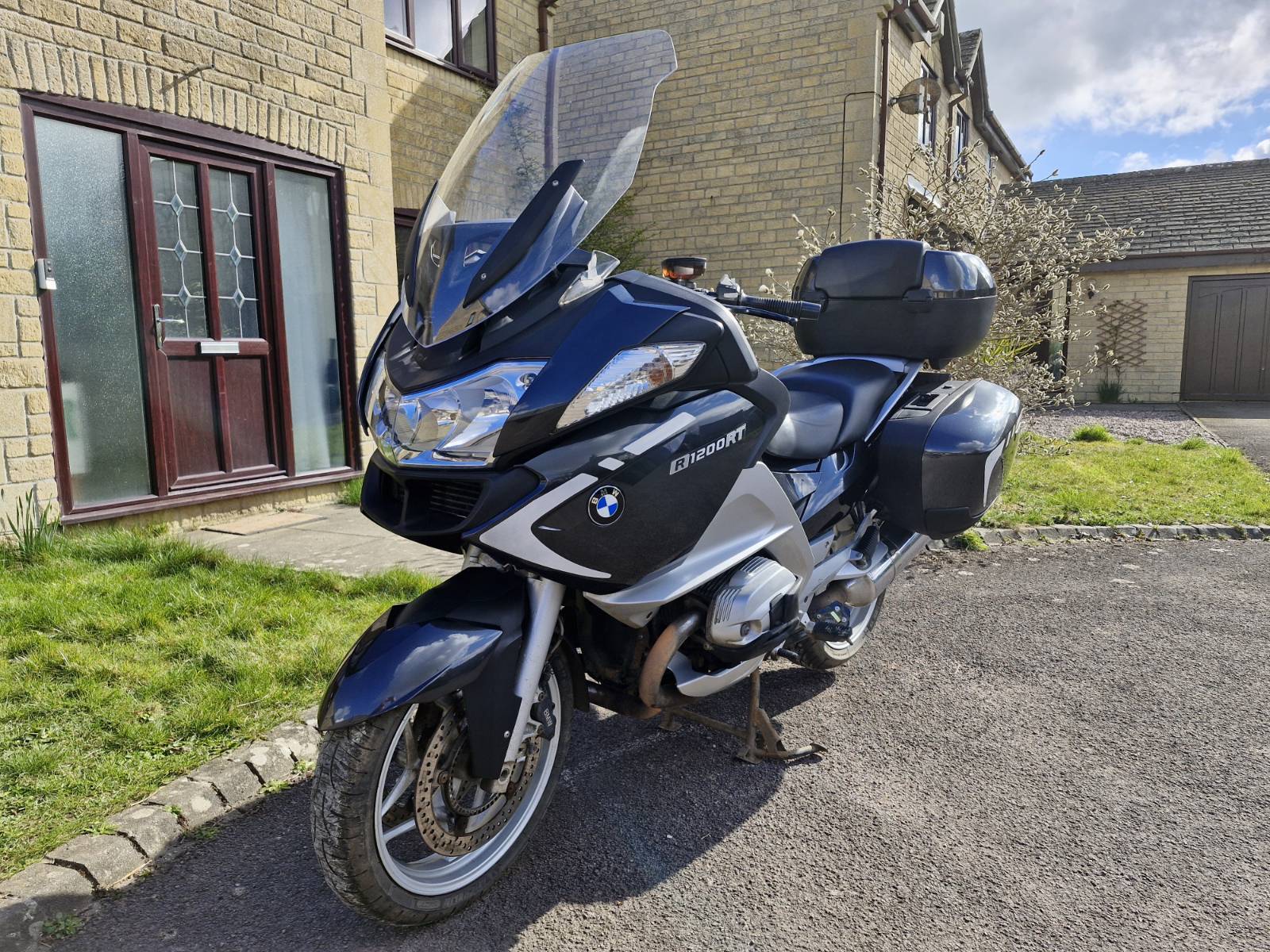 Image features a BMW R1200RT motorcycle parked on a driveway in front of a stone house. The motorcycle is styled with a sleek, aerodynamic design, predominantly in black with silver accents. The front fairing includes a large, clear windshield, and the BMW logo is prominently displayed on the side. The bike is equipped with side panniers and a top case for storage.